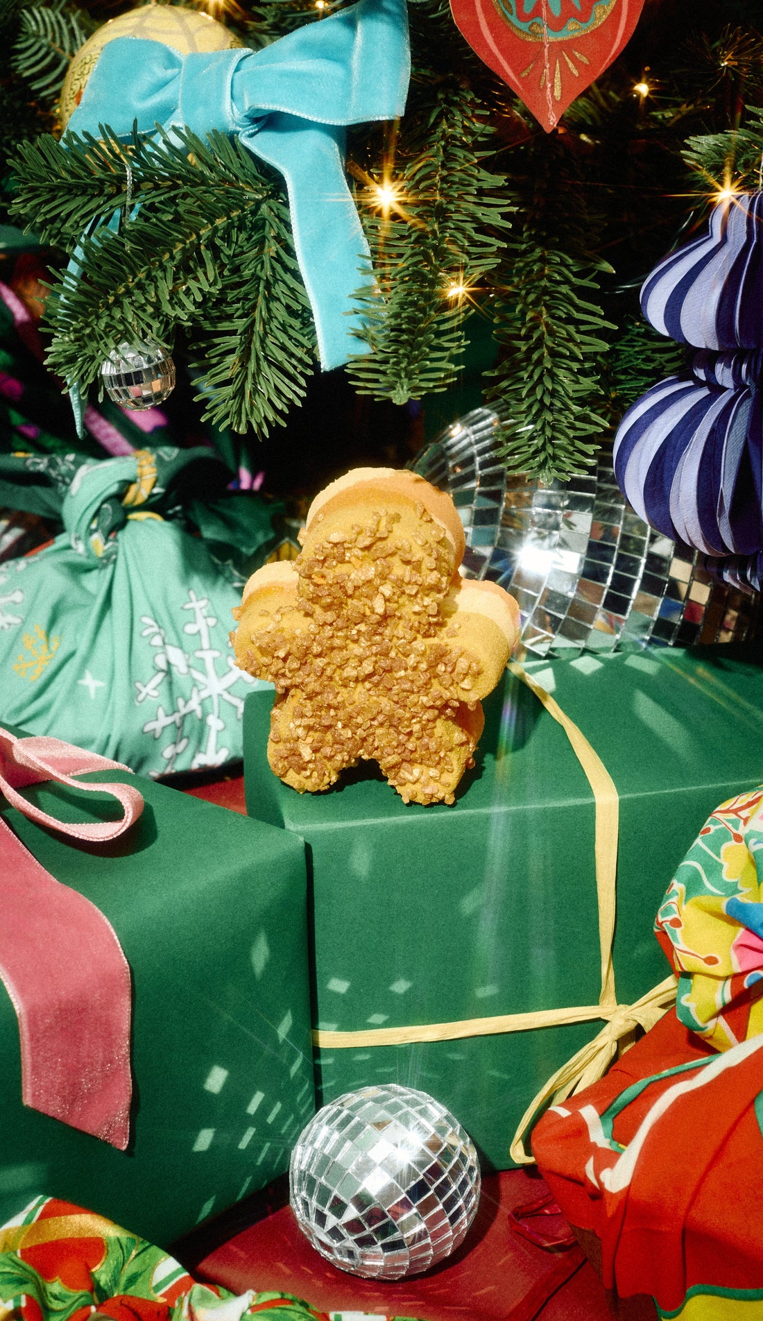 Gingerbread Sparkle gingerbread-shaped bath bomb resting on a green present beneath a Christmas tree.