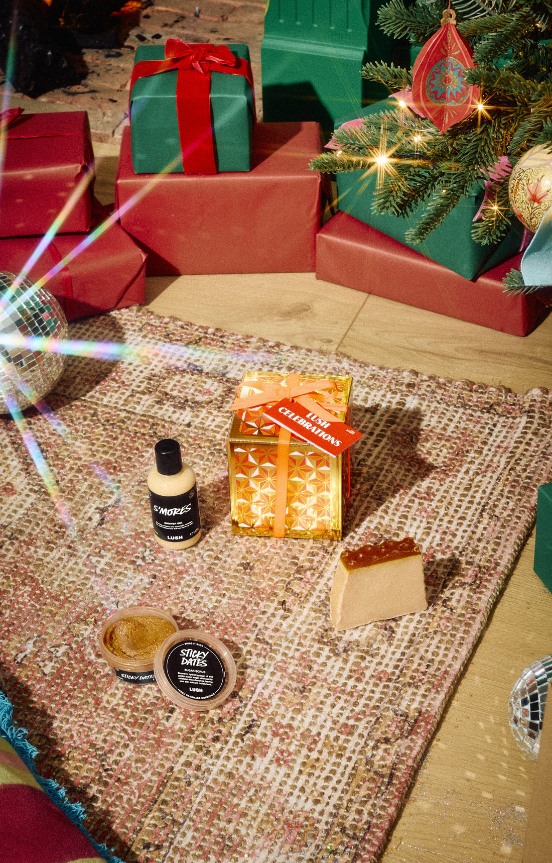 Lush Celebrations gold gift box displayed with a bottle, open pot and pale solid product on a rug.