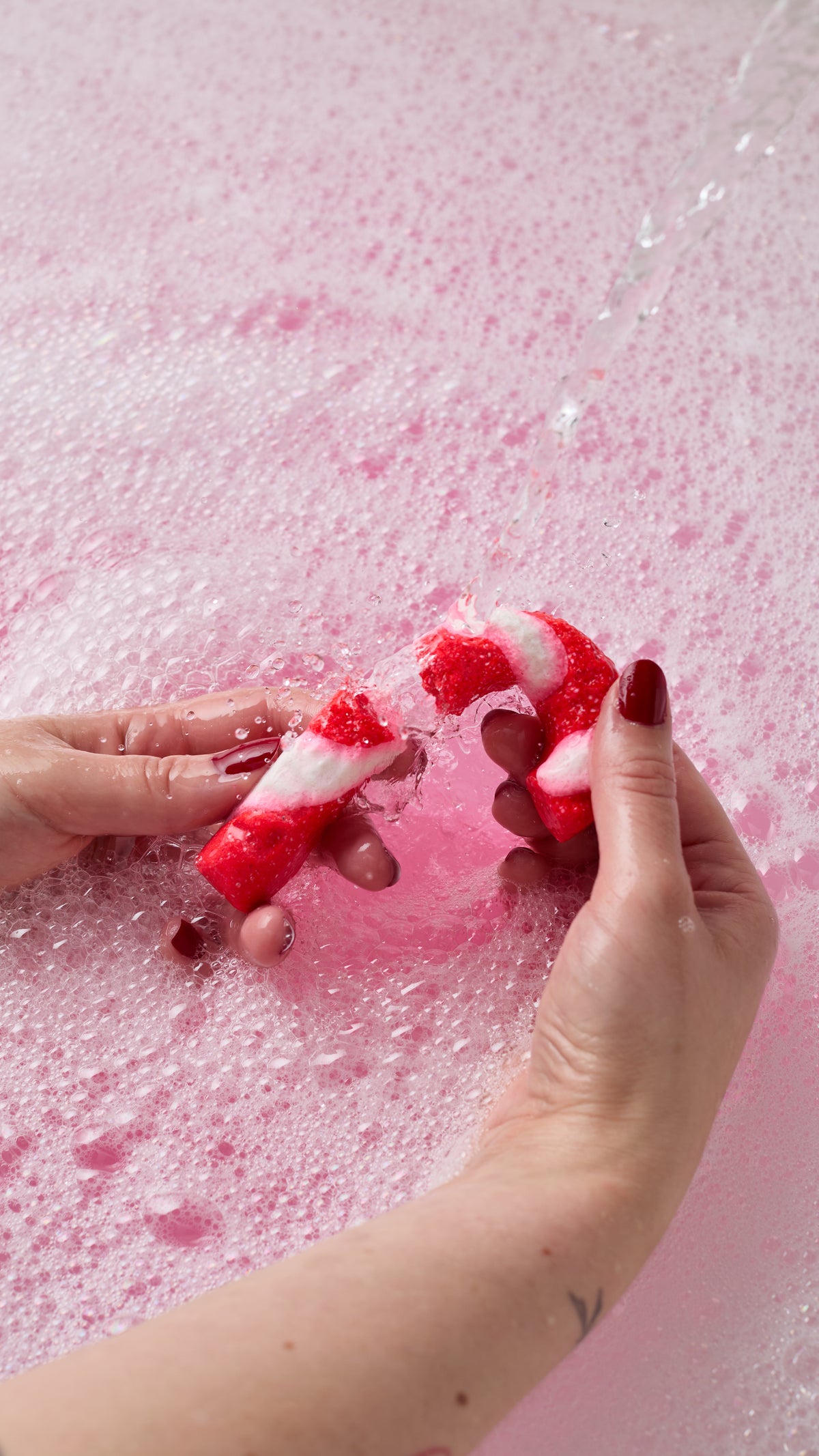 Hands holding Candy Cane bubble bar beneath running water over pink bath water.