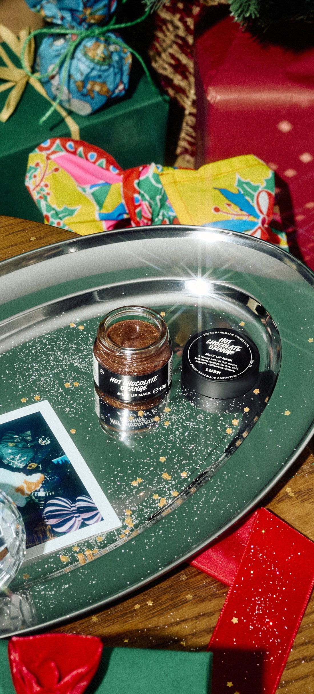 Open Hot Chocolate Orange lip mask jar and black lid on a silver tray among Christmas gifts.