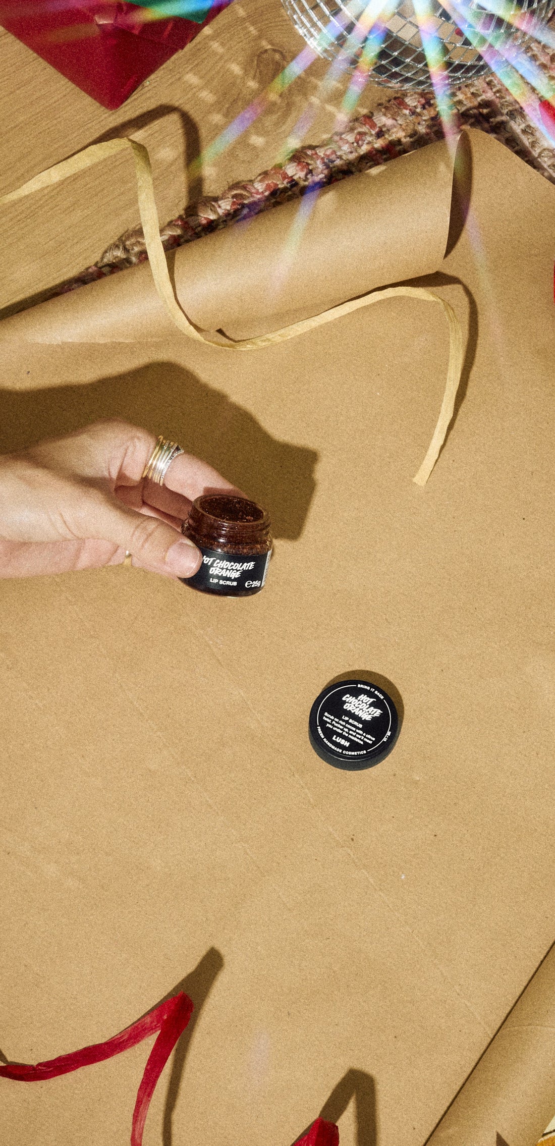 Hand holding an open Hot Chocolate Orange Lip Scrub jar above gold wrapping paper and ribbon.