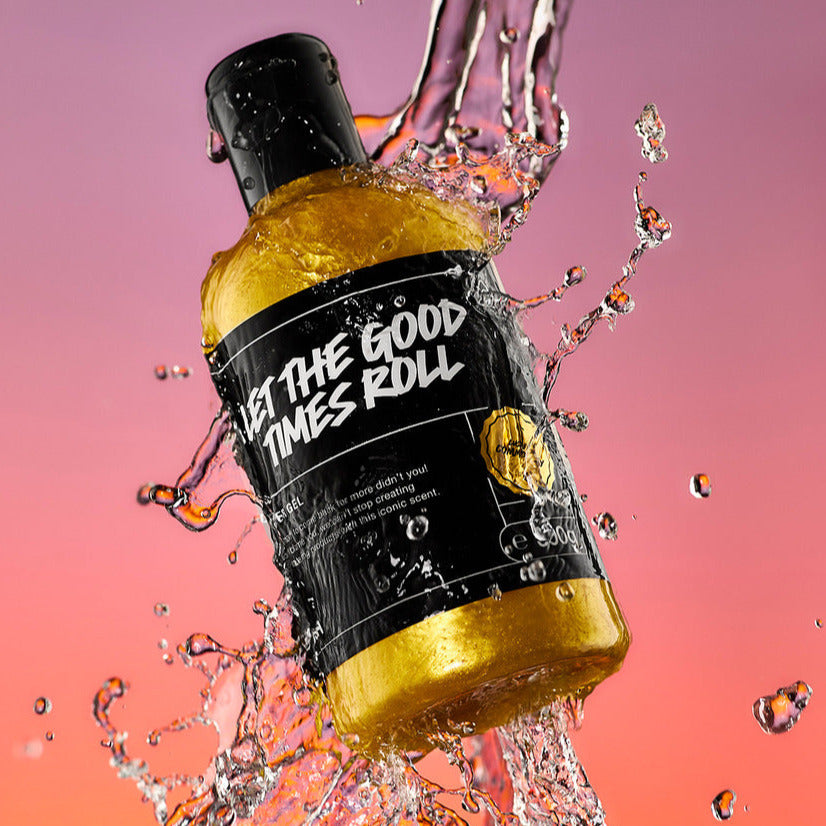 A high-definition action shot of the Let The Good Times Roll shower gel bottle mid-air being splashed by fresh water surrounded by droplets on a blurred purple and orange background.