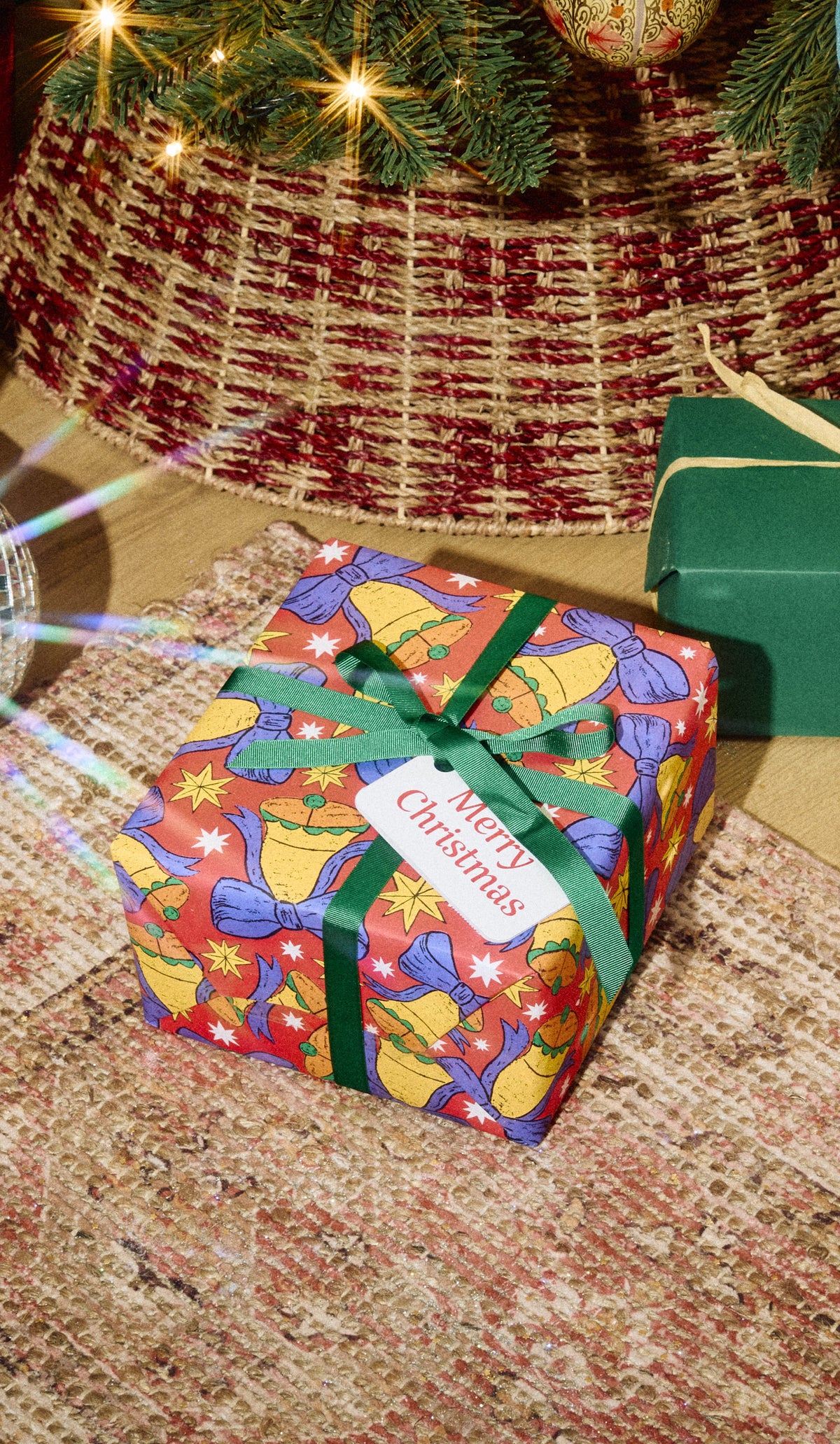 Merry Christmas gift wrapped in colourful festive paper with a green ribbon and white tag beneath a Christmas tree.