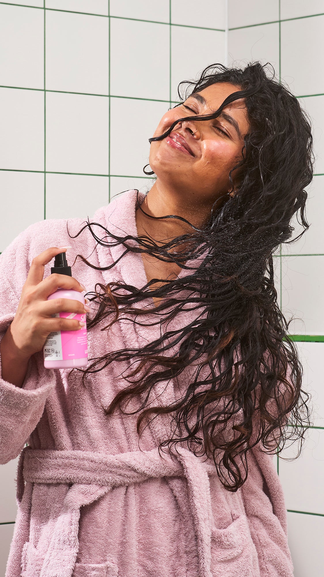 A person in a pink robe holds Snow Fairy hair primer while lifting and separating their curls.