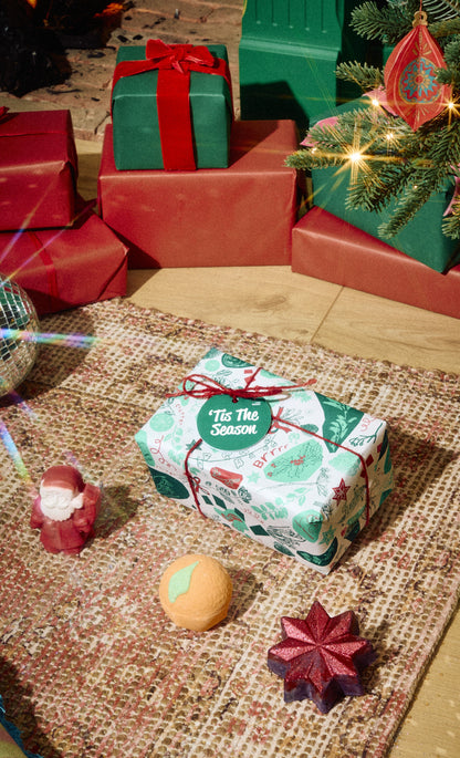 Tis the Season wrapped gift displayed with three bath products on a rug.