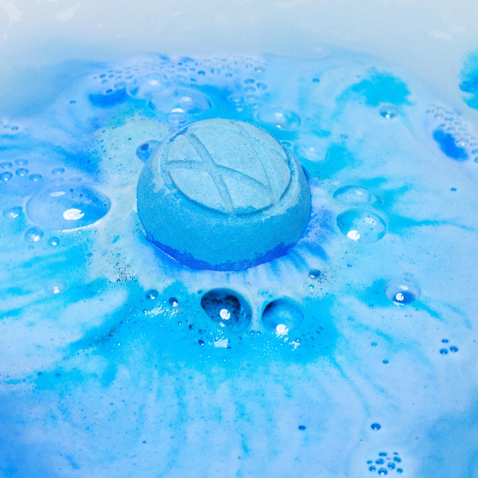 The Hexagon bath bomb is dissolving in the bath water leaving behind ocean-like swirls of deep blues and indigo with lightly foamy bubbles.