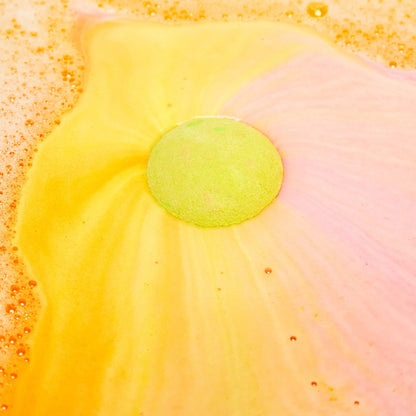 The Time Of The Season bath bomb has been placed into the bath water where it sits like a giant round sun in the centre shooting off swirls of bright yellow and subtle pink like a sunset.