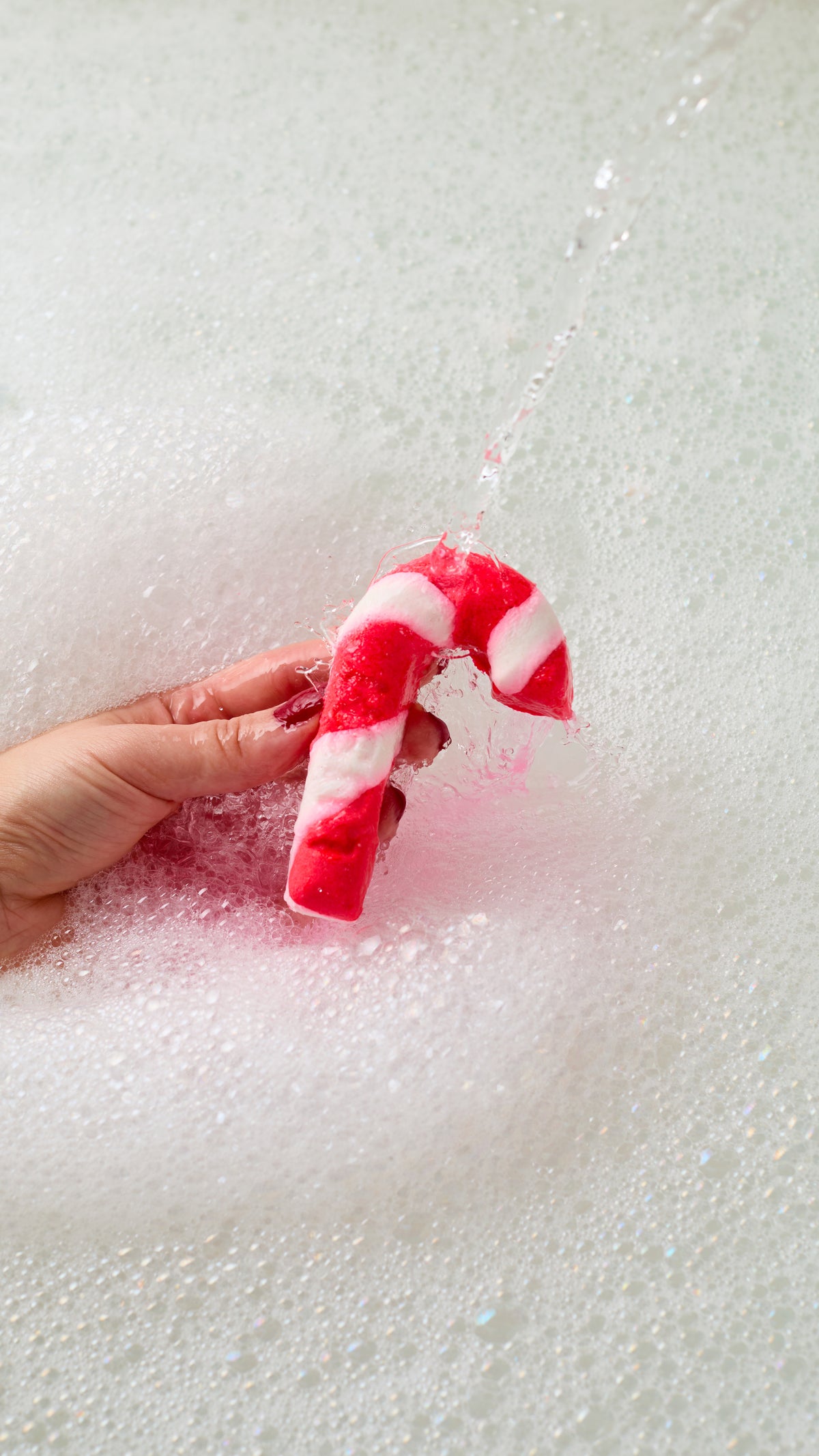 A hand holding Candy Cane bubble bar above white bath bubbles.
