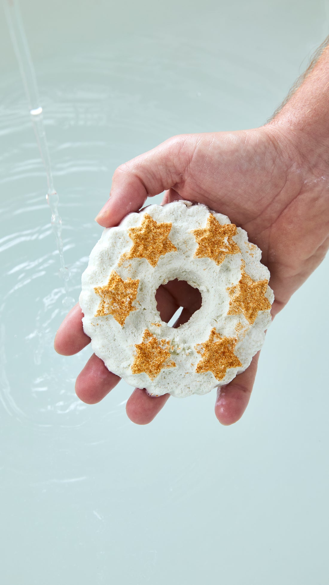 A hand holding the white Christmas Wreath reusable bubble bar with gold stars above bath water.
