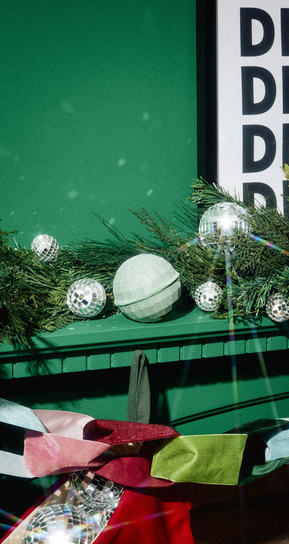 Dancing Shoes pale green round foot soak among evergreen branches on a green mantelpiece.