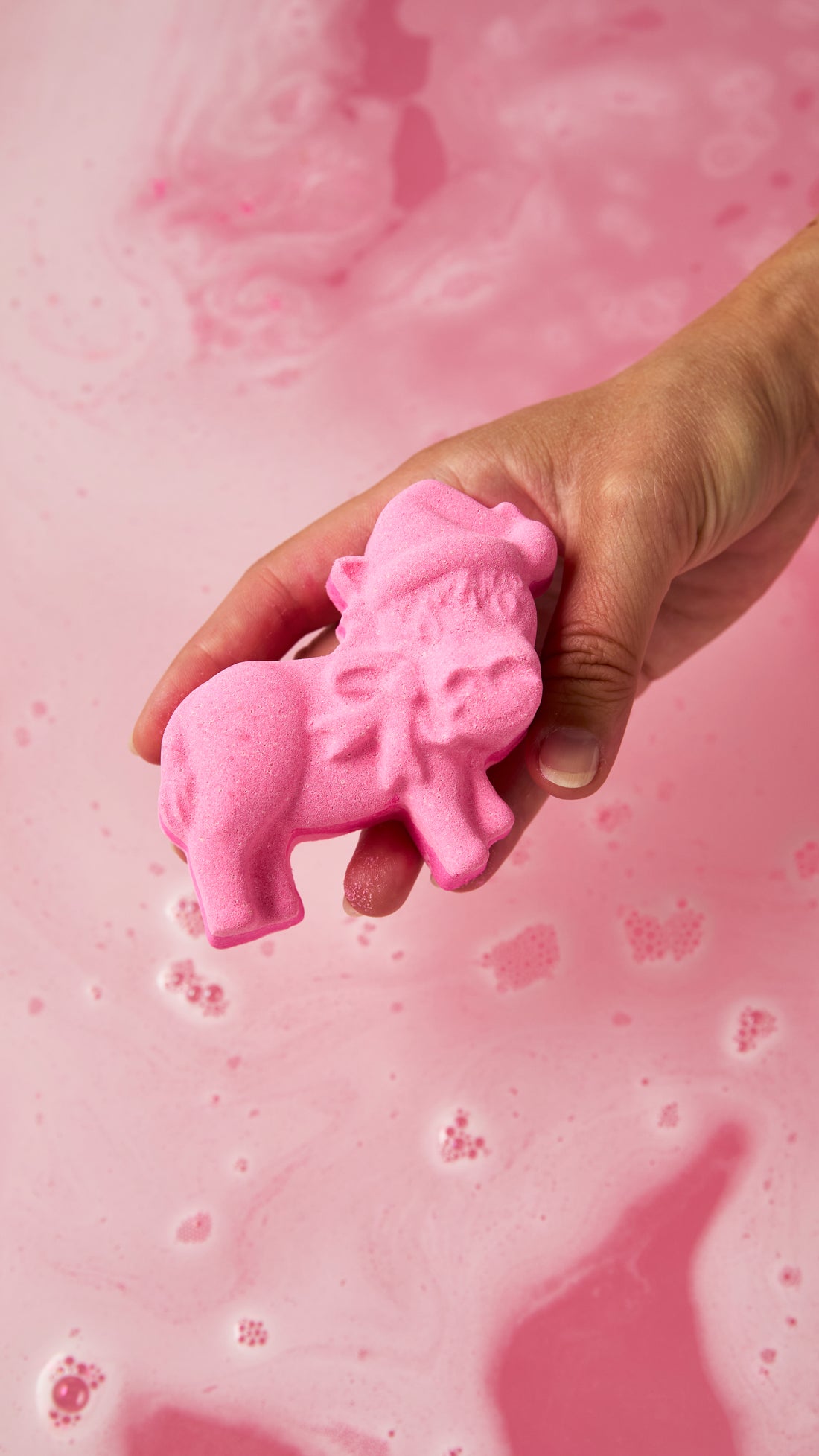 Hand holding the pink donkey-shaped Dominick The Donkey bath bomb above pink bathwater.