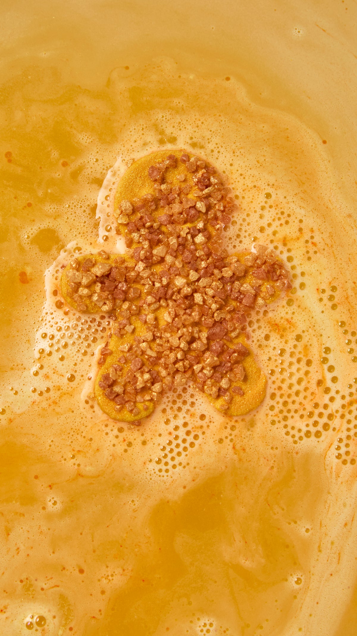 Gingerbread-shaped bath bomb fizzing in golden-orange water with sparkling foam.