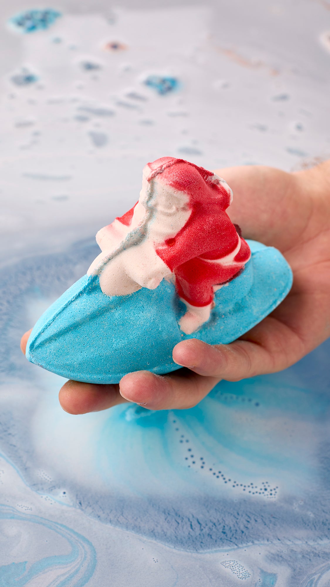 Hand holding the red, white and blue Jet Ski bath bomb over lightly foaming blue water.