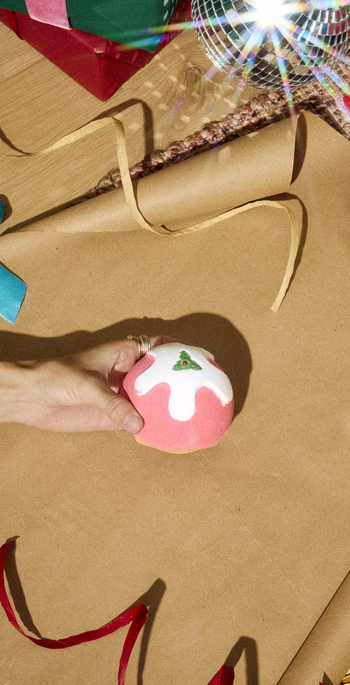 Hand holding the pink Lush Pudding bath bomb with white icing and a holly decoration above gold wrapping paper.