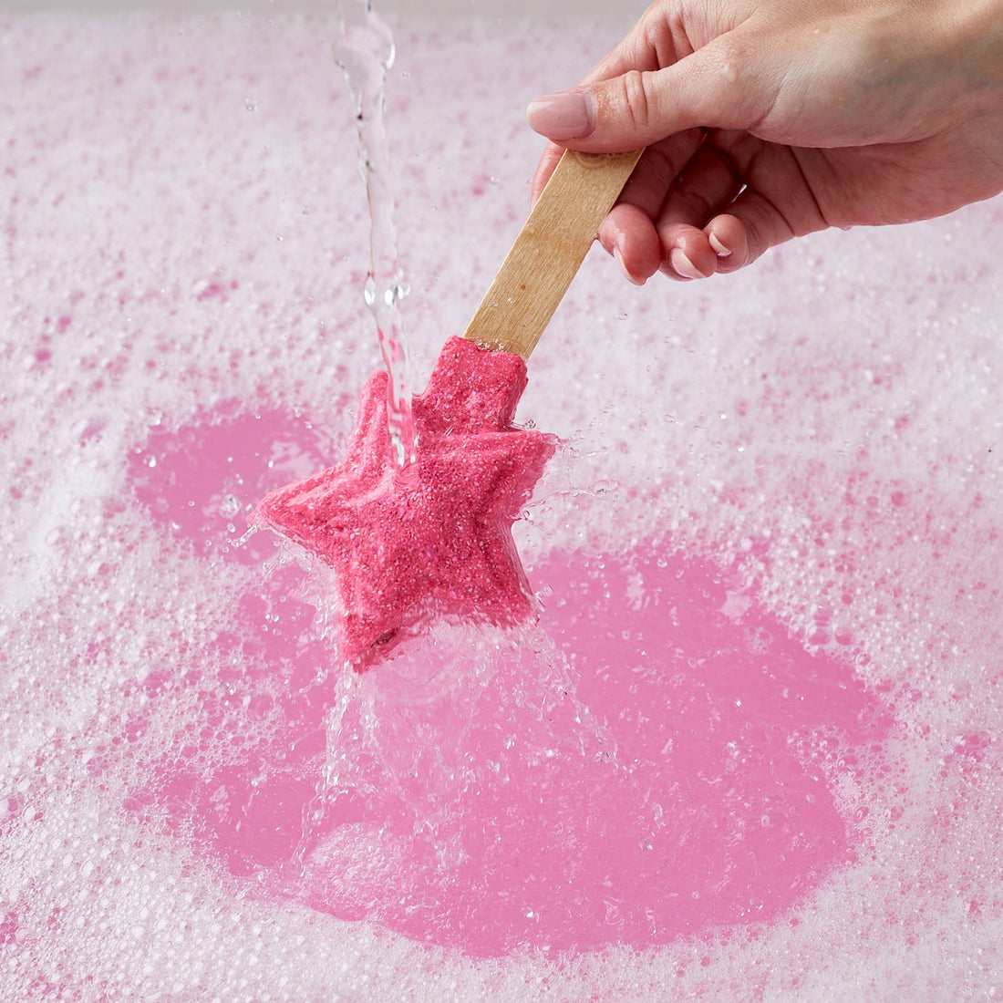 Pink star-shaped bubble bar being dropped into water with a wooden stick, creating bubbles on a light pink background.