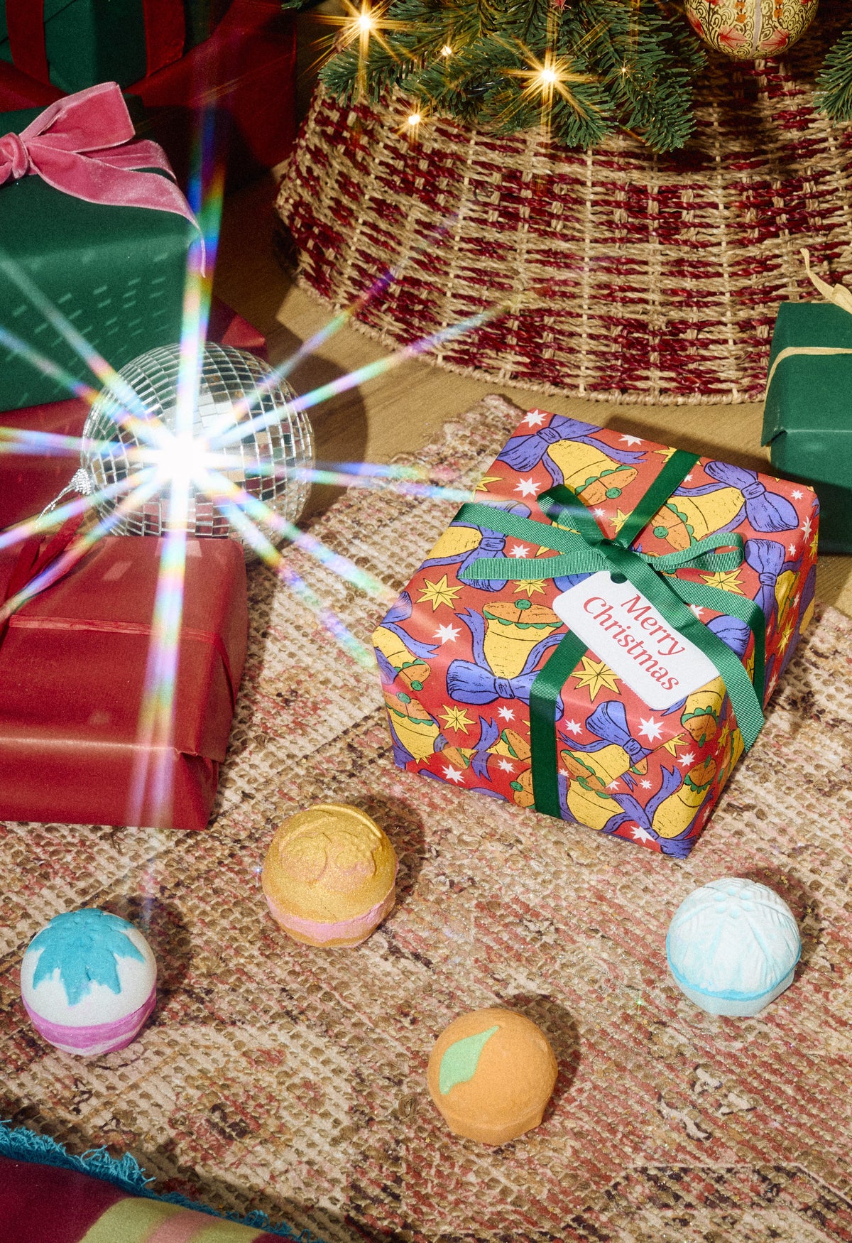 Merry Christmas gift box beside four colourful bath bombs on a rug among wrapped presents.