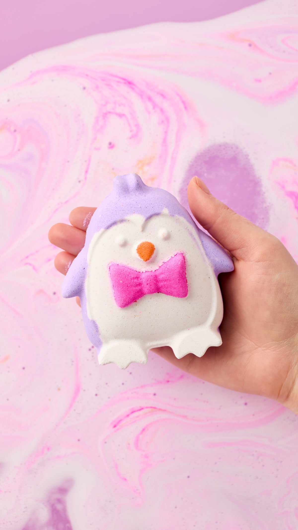 Hand holding The Christmas Penguin bath bomb, with purple body and pink bow tie, above pink and purple bathwater.