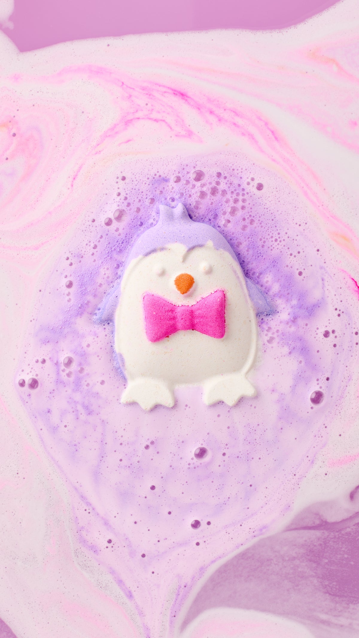 The Christmas Penguin bath bomb floating in lavender foam with pink swirls around it.