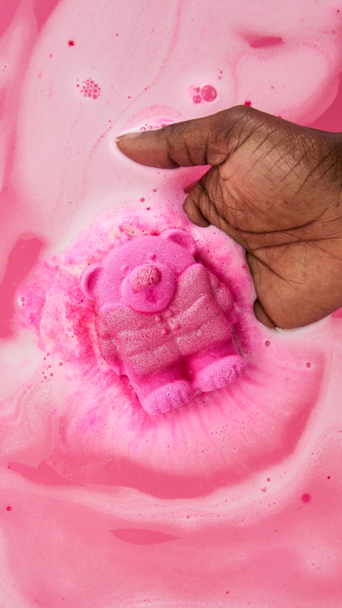 Hand holding a pink bear-shaped bath bomb as it fizzes in bright pink bathwater.
