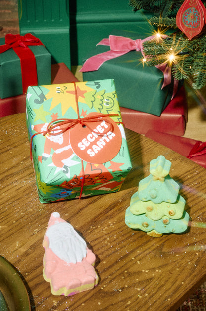Secret Santa gift wrapping beside pink Santa-shaped and green Christmas-tree-shaped bath bombs.