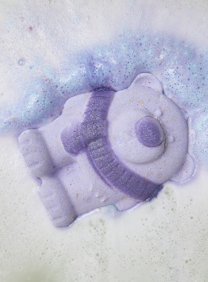 Purple bear-shaped bath bomb dissolving in pale lavender bubbles.