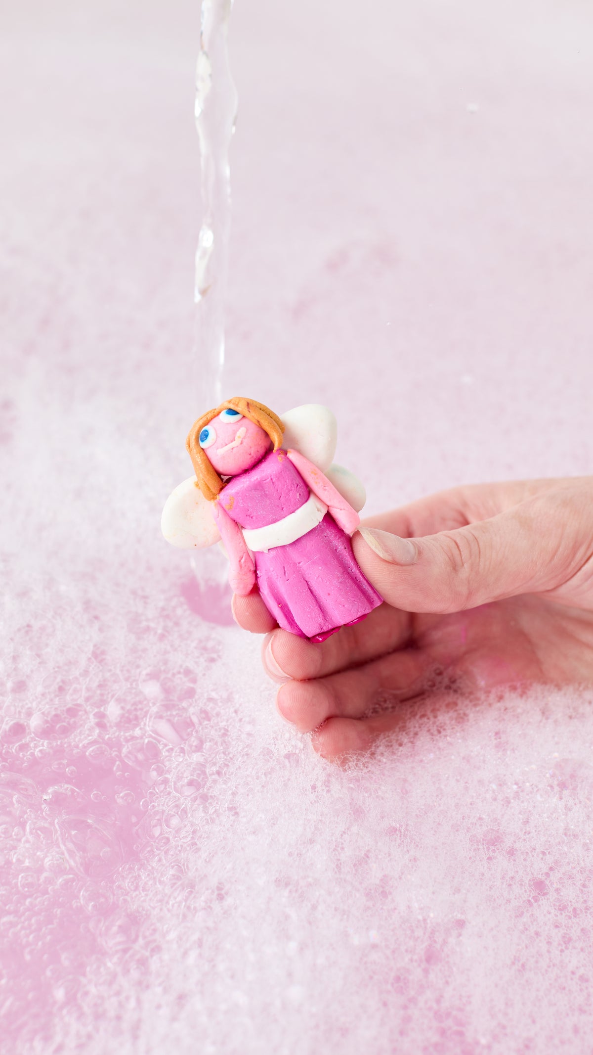 A hand holds Snow Fairy Fun moulded into a pink fairy figure beneath running bathwater.