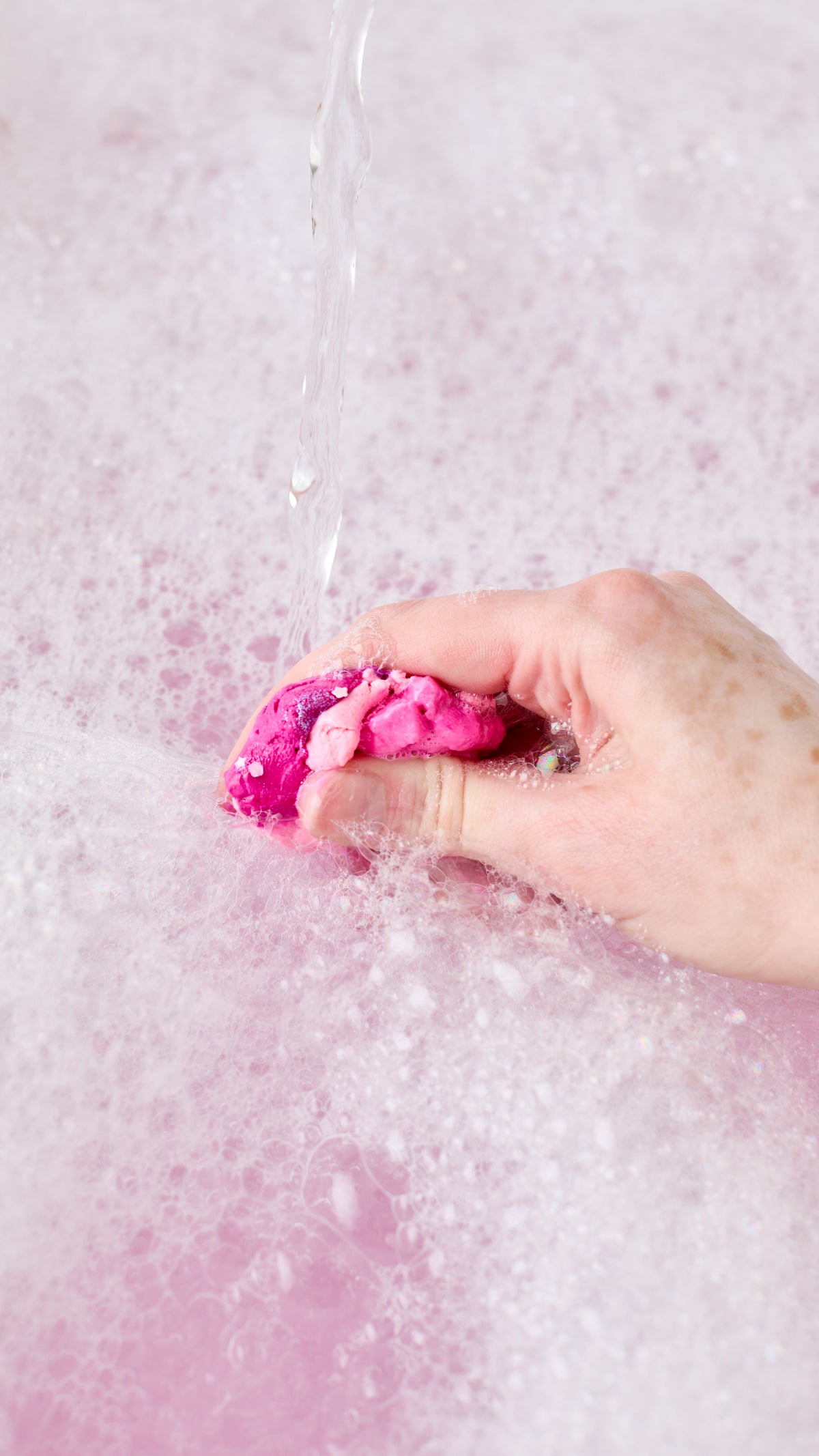 A hand squeezes pink Snow Fairy Fun beneath running water in a foamy bath.