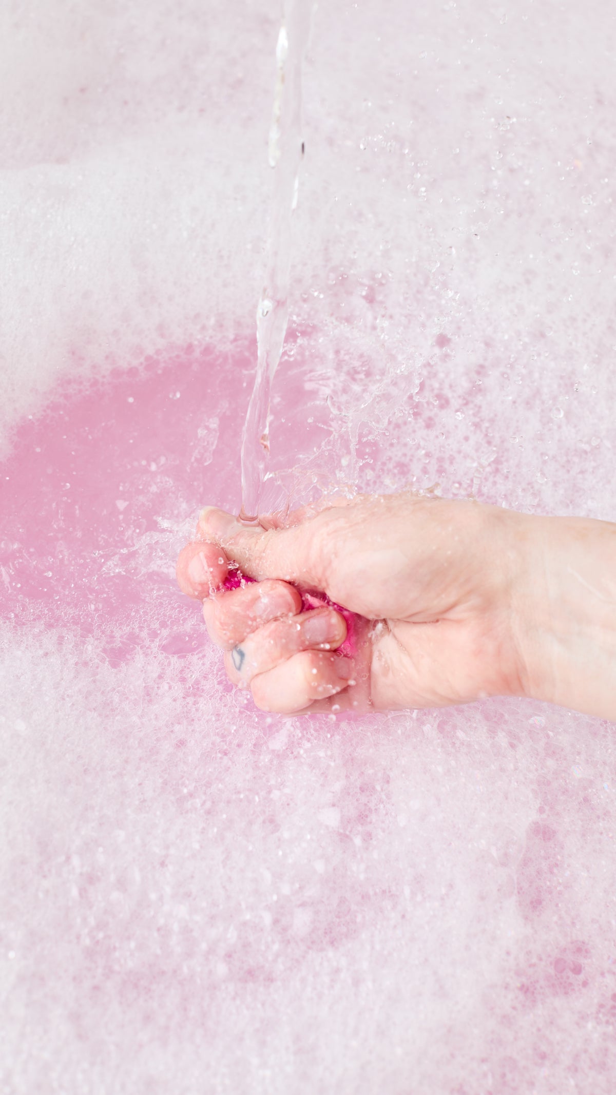 A hand crumbles Snow Fairy Fun beneath running water, creating pink bathwater and bubbles.