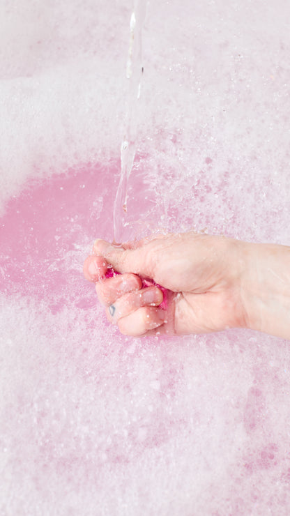 A hand crumbles Snow Fairy Fun beneath running water, creating pink bathwater and bubbles.