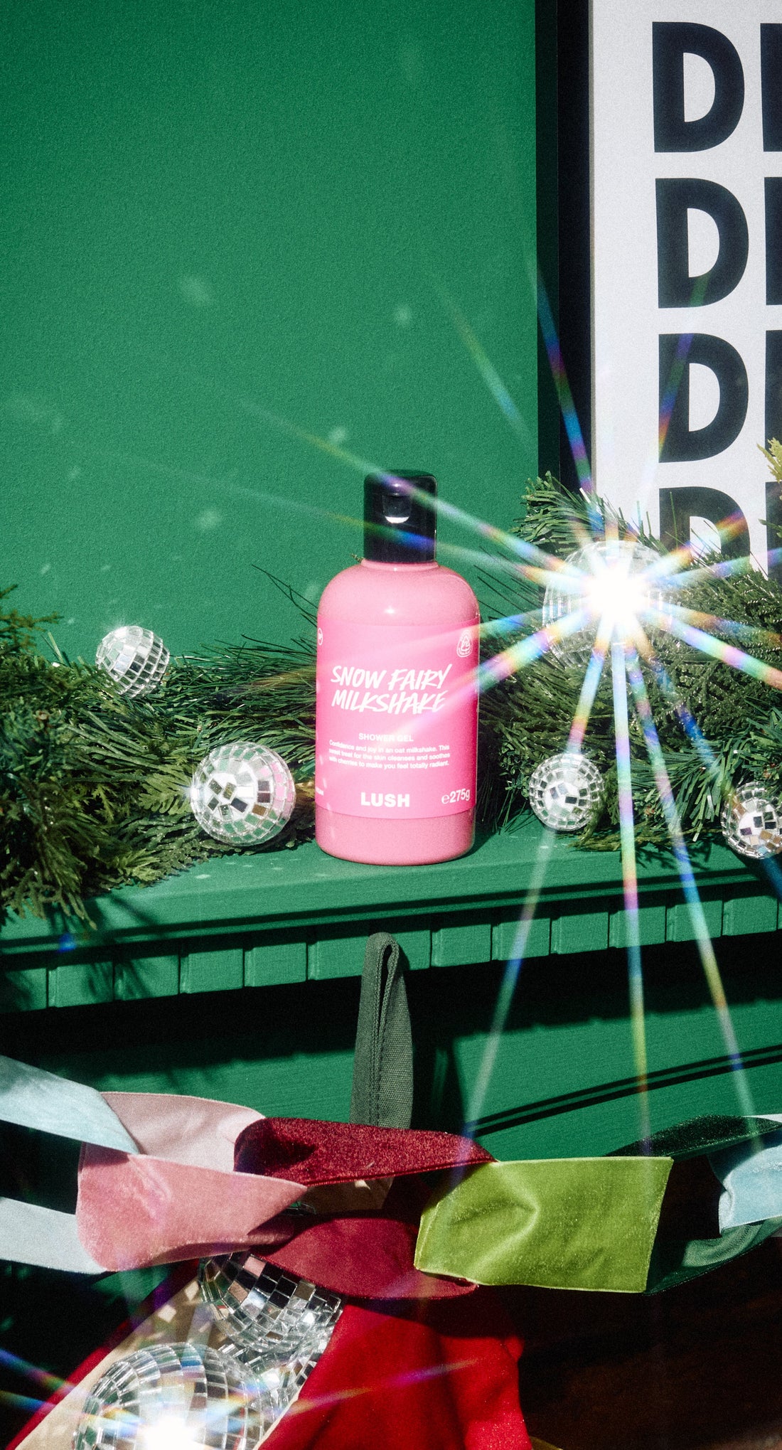 A pink Snow Fairy Milkshake shower gel bottle stands on a green mantel among silver baubles and festive foliage.
