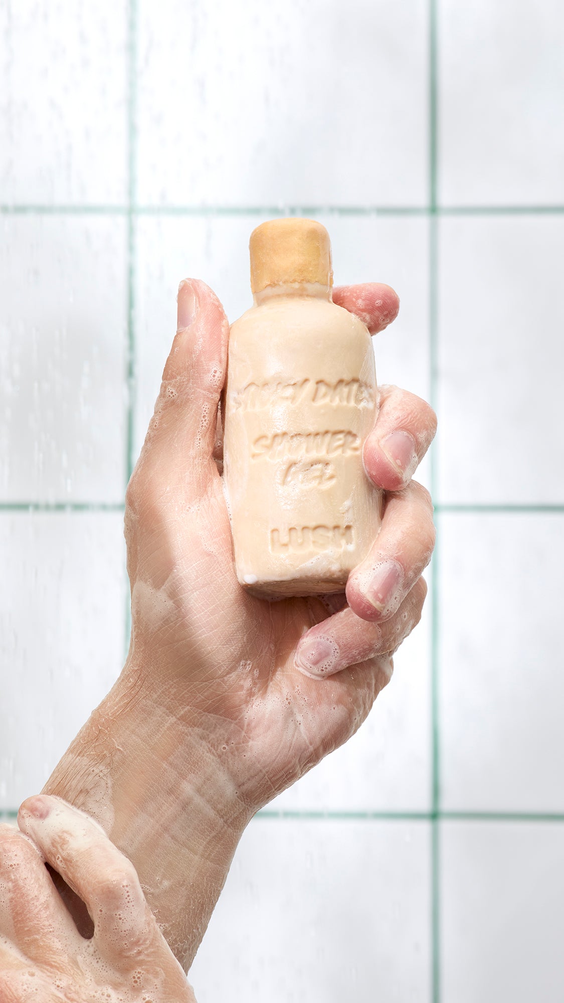 A hand holds tan, bottle-shaped Sticky Dates naked shower gel against bathroom tiles.