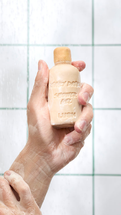 A hand holds tan, bottle-shaped Sticky Dates naked shower gel against bathroom tiles.