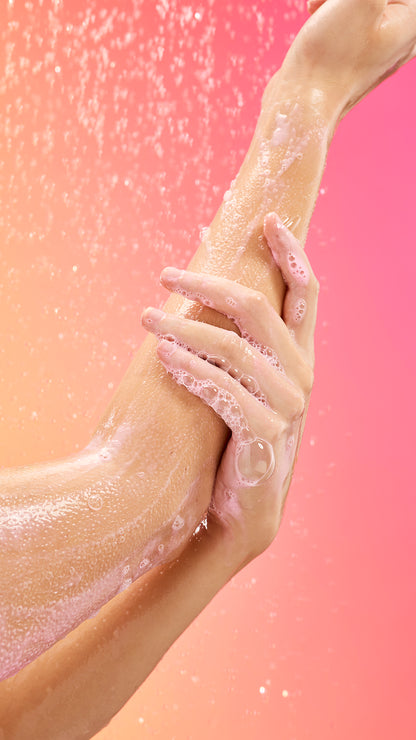 A hand massages pink body scrub along a wet forearm against a bright pink background.