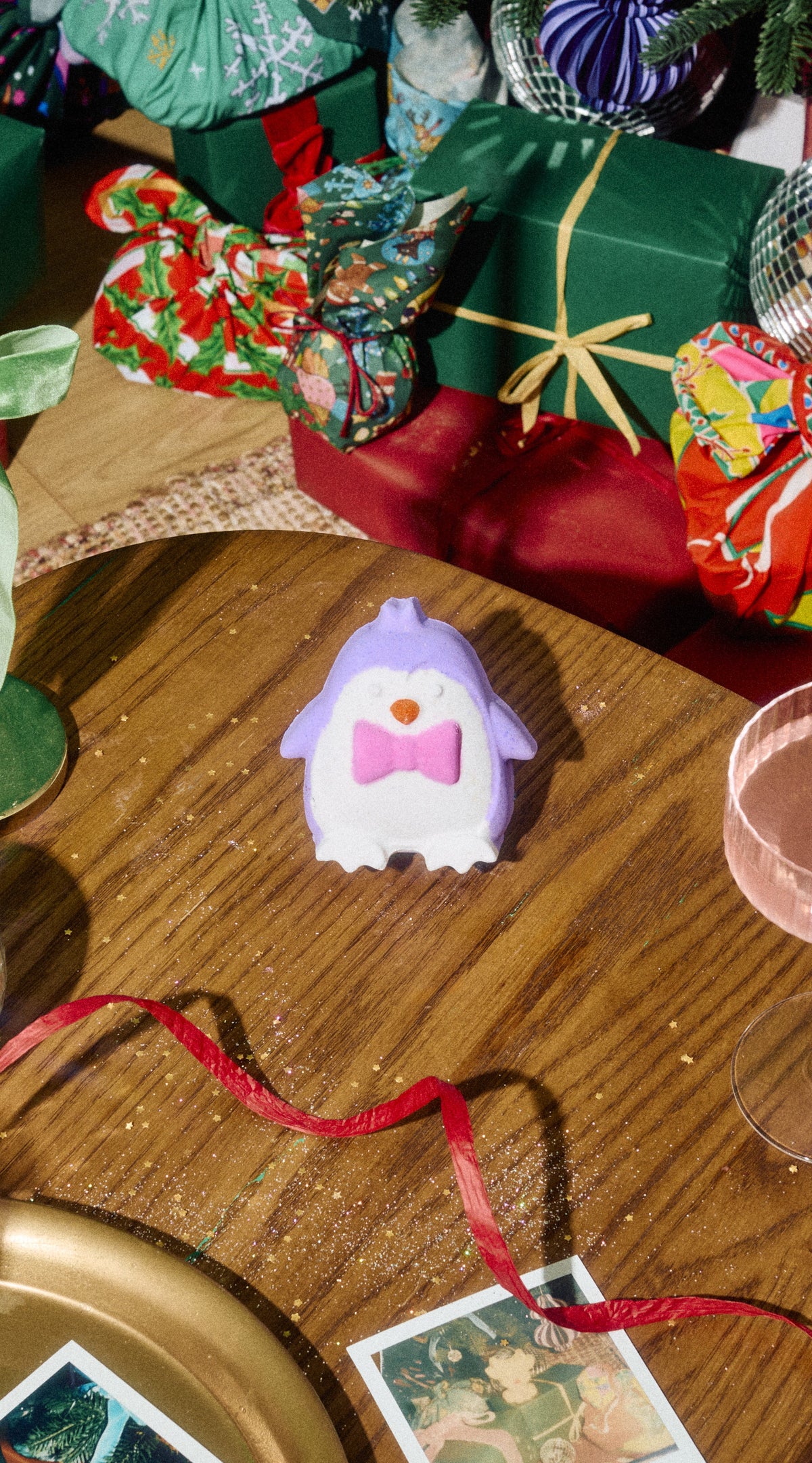 Purple and white Christmas Penguin bath bomb with a pink bow tie on a table among festive presents.