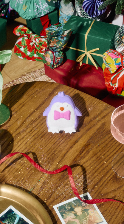 Purple and white Christmas Penguin bath bomb with a pink bow tie on a table among festive presents.