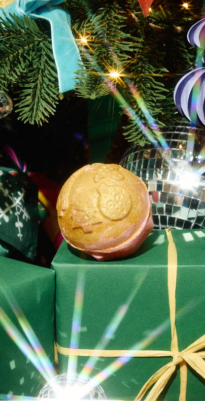 A round golden Yog Nog bath bomb displayed on a green gift box beneath a Christmas tree.